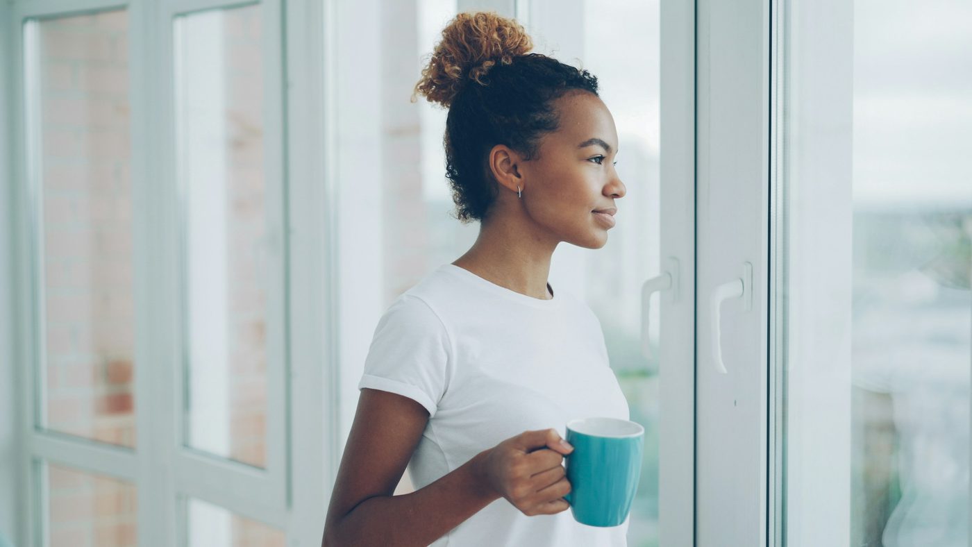 A woman standing at a large window with a mug of tea, looking out at the daylight